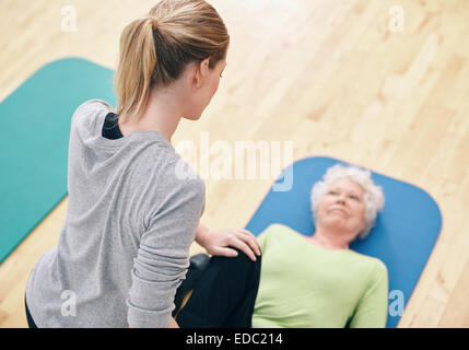 Vista ad angolo alto di una coach che aiuta una donna anziana ad allenarsi in palestra. allenatrice di palestra che aiuta una donna anziana nel lavoro di stretching delle gambe Foto Stock