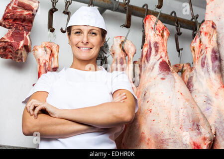 Macellaio in piedi contro la carne appesi in macelleria Foto Stock