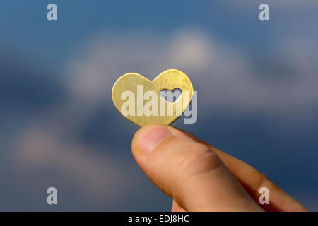 Una mano è in possesso di una bella luminosa forma di cuore con un piccolo cuore in esso, sullo sfondo di un cielo blu con nuvole. Foto Stock