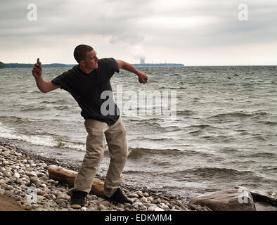 Giovane uomo saltando pietre sul Lago Ontario, New York . Nine Mile punto di generazione nucleare impianto è nella distanza Foto Stock