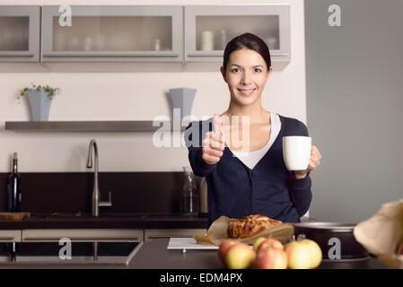 Felice attraente femmina giovane cuoco dando un pollice in alto di approvazione e di successo come lei sta nella sua cucina a bere caffè Foto Stock