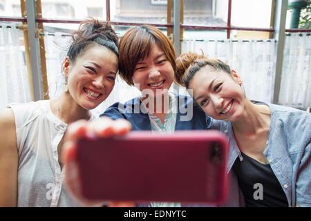 Tre donne alla ricerca di un telefono cellulare, prendendo un selfie, seduta all'interno. Foto Stock