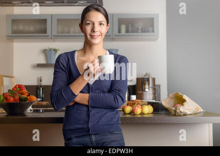 Felice contenti casalinga nella sua cucina dando la telecamera un caldo sorriso amichevole come lei si rilassa con una tazza di caffè con freschi Foto Stock