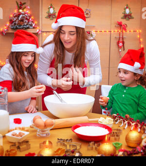 Felice madre di due bambini carino indossando red Santa hat cucinare cibo festoso sulla cucina, vacanze di Natale del concetto Foto Stock