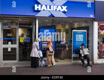 Due acquirenti femmina usando un bancomat e un suonatore ambulante di fisarmonica al di fuori Halifax UK bank in Walsall, West Midlands, Inghilterra. Foto Stock