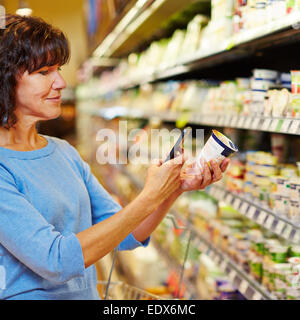 Donna anziana con smartphone codice a barre a scansione di yogurt in un supermercato Foto Stock