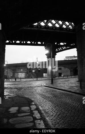 Vista serale in bianco e nero attraverso un arco ferroviario in ferro vittoriano su ciottoli bagnati ed edifici industriali in mattoni nel centro di Manchester. Foto Stock