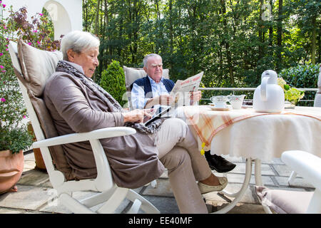 Coppia senior, metà 70, a mezzogiorno, di bere il caffè sulla terrazza, Lei naviga con un tablet PC, iPad Internet, la lettura di un newspa Foto Stock