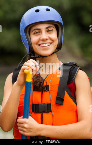 Una giovane donna che indossa un casco blu e un giubbotto di salvataggio arancione sorride alla fotocamera mentre tiene in mano una pagaia blu e gialla. Sembra essere all'aperto, probabilmente vicino all'acqua, prepararsi o fare una pausa Foto Stock