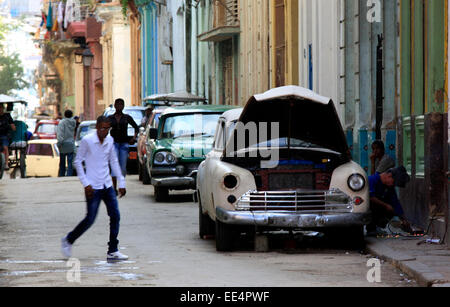 Scena di strada in Avana con vintage American cars parcheggiato sulla strada e un uomo di riparazioni di saldatura per la sua auto crema Foto Stock