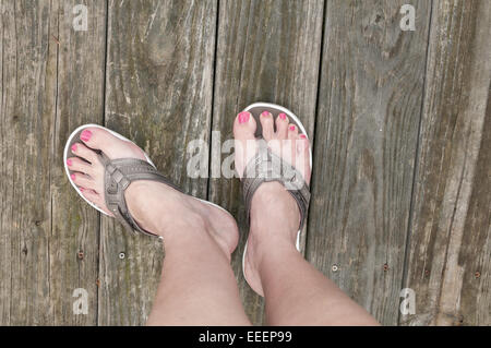 I piedi femminili in piedi su un ponte di legno in una coppia di color bronzo di Sandali e infradito. Foto Stock