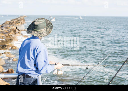 L'uomo con il cappello per il sole visto dal retro in piedi in una ringhiera con canna da pesca in mano che dava sull'oceano, pietra jetty e barche. Foto Stock