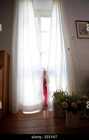 Ragazza giovane di giocare a nascondino dietro bianco opaco, tende di fronte ad una grande finestra in una casa.stile di vita Foto Stock