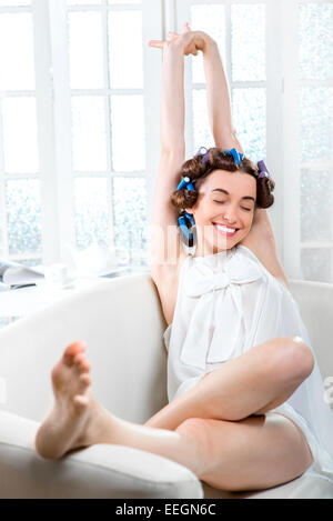 Giovane donna con bigodini in her hair sbadigliare e giacente nel lettino al bianco interni domestici Foto Stock