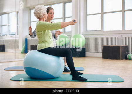 Trainer femmina assistere senior donna sollevamento pesi nella palestra. Senior donna seduta sulla sfera di pilates facendo esercizio di peso. Foto Stock