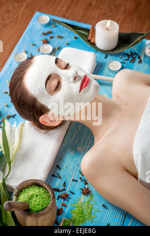 Giovane donna con centro termale viso maschera sul suo viso giacente sul tavolo blu con fiori, candele e sale marino in beaty salon Foto Stock