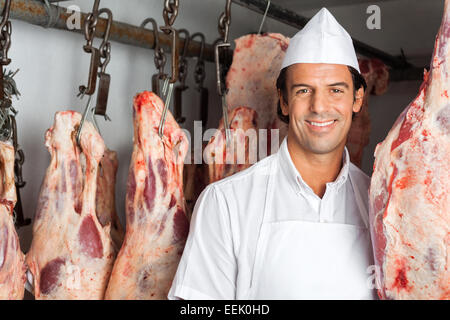 Macellaio in piedi vicino a carne appesi in macelleria Foto Stock