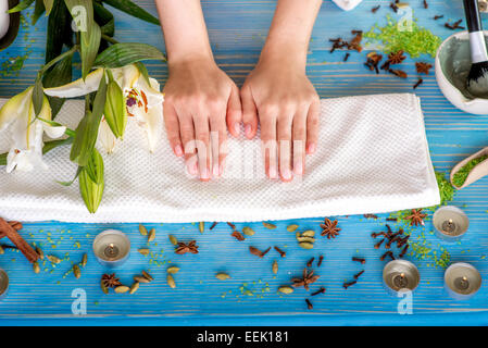 Donna prendendo cura delle sue mani sul telo da bagno in spa con fiori, candele e erbe blu sulla scrivania in legno Foto Stock