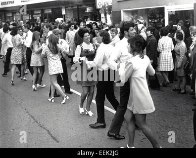 Anni sessanta storici, gli acquirenti la visione di uomini e donne che danzano insieme in strada con le ragazze che indossano floreali e abiti breve mini gonne, Londra, Inghilterra. Possiblly Carnaby Street, il centro della moda inglese in questa epoca. Foto Stock