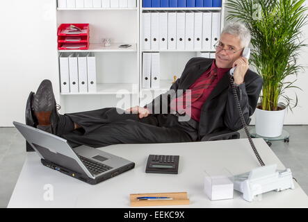 Mann mittleren altera sitzt im Büro hat lässig die Beine auf dem Schreibtisch und telefoniert, uomo di mezza età si siede in un ufficio Foto Stock