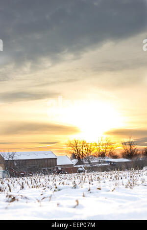 Inverno del racconto. Paesaggio invernale innevato con il villaggio di campagna accanto a cornfield coperto di neve bianca coprire al tramonto o sunris Foto Stock