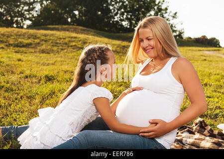 Carino bambina abbracciando la madre del ventre in gravidanza. Felice la sorella in essere è entusiasta di incontrare nuove baby. La festa della mamma concept Foto Stock