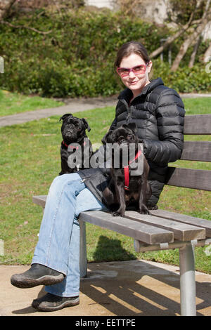 Donna nella sua anni trenta seduti su un parco urbano banco con le sue due Pugs nero, fagiolo e Frank Foto Stock