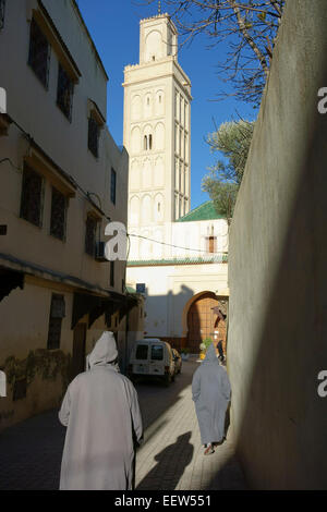 Gli uomini marocchini a piedi indossando il tradizionale djellaba incappucciato, minareto della moschea Berdaine, Meknes, Marocco Foto Stock