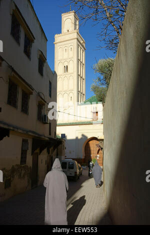 Gli uomini marocchini a piedi indossando il tradizionale djellaba incappucciato, minareto della moschea Berdaine, Meknes, Marocco Foto Stock