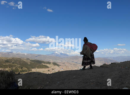 Una donna indigena in abito tradizionale passeggiate contro lo sfondo delle Ande e la Bolivia è la seconda montagna più alta, Illimani. Foto Stock
