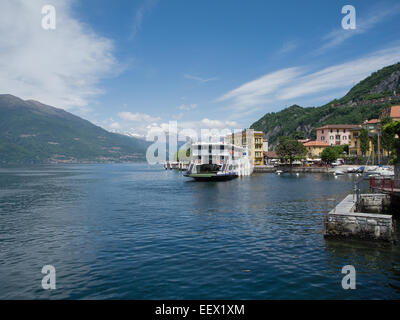 Varenna sul Lago di Como in Italia con un traghetto che sta per lasciare il molo e uno sfondo di montagne innevate e un cielo blu Foto Stock