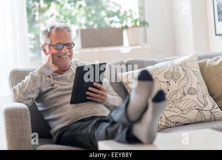 Senior uomo seduto sul divano nel soggiorno, utilizzando un tablet pc e sorridente Foto Stock