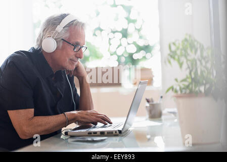 Senior uomo seduto in casa ufficio indossando le cuffie e utilizzando computer portatile Foto Stock