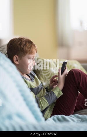 Ragazzo (6-7) seduto sul divano e utilizzando il telefono cellulare Foto Stock