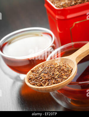 Rooibos in cucchiaio di legno closeup, due tazze di tè e tè la scatola di stagno su tavola Foto Stock