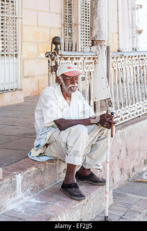 Gentile ma gli infermi locale di vecchio uomo vestito di bianco vestiti con un bastone da passeggio seduta sui gradini di fumare un sigaro cubano nel centro cittadino di Trinidad, Cuba Foto Stock