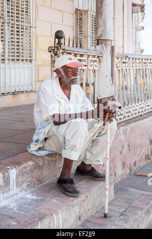 Gentile ma gli infermi locale di vecchio uomo vestito di bianco vestiti con un bastone da passeggio seduta sui gradini di fumare un sigaro cubano nel centro cittadino di Trinidad, Cuba Foto Stock