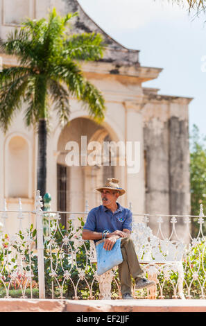 Stile di vita cubana locale: uomo di mezza età con un baffi indossando un cappello di paglia rilassante seduta su una panchina di fumare un sigaro cubano, centro di Trinidad, Cuba Foto Stock