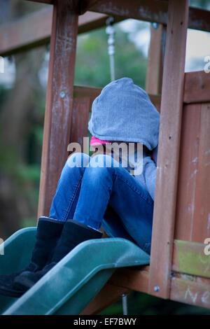 Triste bambino in felpa con cappuccio con la faccia nascosta in mani, seduto da solo in un parco giochi nella disperazione. Foto Stock