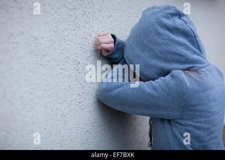 Triste bambino in felpa con cappuccio con la faccia nascosta, appoggiato contro una parete nella disperazione Foto Stock