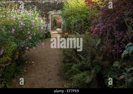 Un ampio sentiero di ghiaia conduce ad un arco di pietra, fiancheggiata da Herbaceous borders, alla fine di ottobre, Rousham House, Oxfordshire, Inghilterra Foto Stock