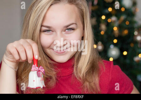 Ritratto di una ragazza adolescente sorridente davanti all'albero di Natale con un ornamento di campanella di Natale Foto Stock