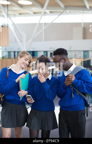 Tre studenti felici che indossano uniformi scolastiche utilizzando lo smartphone nel corridoio della scuola Foto Stock