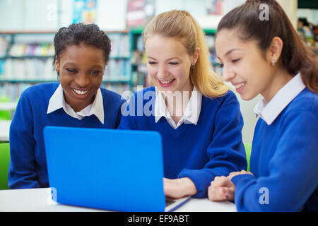 Tre sorridente studenti femmina blu che indossano uniformi scolastiche lavorando sul computer portatile in biblioteca Foto Stock
