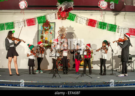 Rifiniti in cappelli di Babbo Natale, Ispanica giovani studenti di musica riprodurre i violini in un concerto di Natale in Santa Ana CA "barrio" centro comunitario auditorium. Nota volontario americano asiatico di alta scuola di musica le guide e il messicano i colori nazionali. Foto Stock