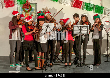 Rifiniti in cappelli di Babbo Natale, Ispanica giovani studenti di musica riprodurre i violini in un concerto di Natale in Santa Ana CA "barrio" centro comunitario auditorium. Nota volontario americano asiatico di alta scuola di musica le guide e il messicano i colori nazionali. Foto Stock