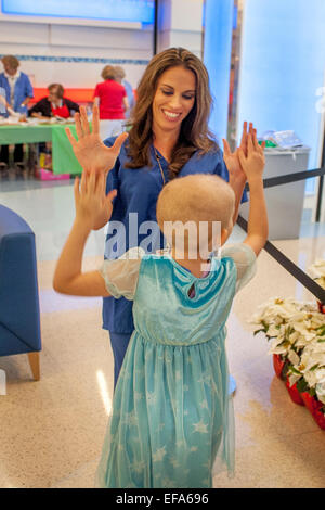 Un bambino sorridente vita specializzato di choc ospedale per bambini in arancione, CA, dà un elevato da cinque a una giovane ragazza in chemioterapia paziente di cancro. Nota testa calva. La vita del bambino il supporto di specialisti i pazienti e le loro famiglie in ambiente ospedaliero . Foto Stock