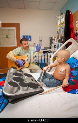 Indossa un abito di sterile e guanti di gomma, una musica terapista di suonare una chitarra suona un giovane ragazzo cancro Chemioterapia paziente a choc ospedale per bambini in arancione, CA. Nota bambino che gioca su tamburi e testa calva. Foto Stock