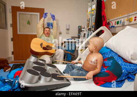 Indossa un abito di sterile e guanti di gomma, una musica terapista di suonare una chitarra suona un giovane ragazzo cancro Chemioterapia paziente a choc ospedale per bambini in arancione, CA. Nota bambino che gioca su tamburi e testa calva. Foto Stock