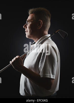 Vista laterale, uomo maturo golfista che indossa una camicia bianca e detiene un ferro golf club sulla sua spalla - studio shot, sfondo nero Foto Stock